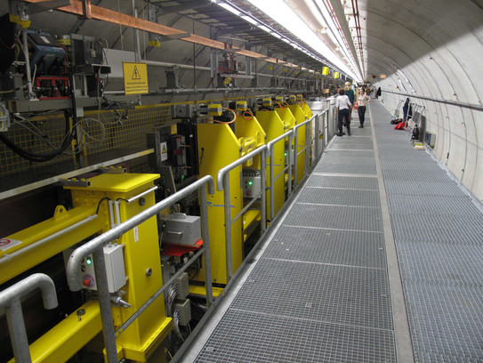Lange Reihe von Undulatoren des Freie-Elektronen-Lasers FLASH in einem Tunnel, im Hintergrund drei Personen.