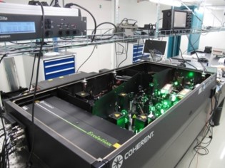 Lasersystem in einem geöffneten schwarzen Kasten. Man sieht viele optische Element und das grüne Licht des Pumplasers. 